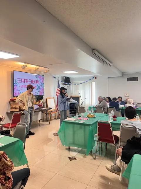 生日會開場,工作人員在螢幕前主持,住戶圍坐綠色餐桌